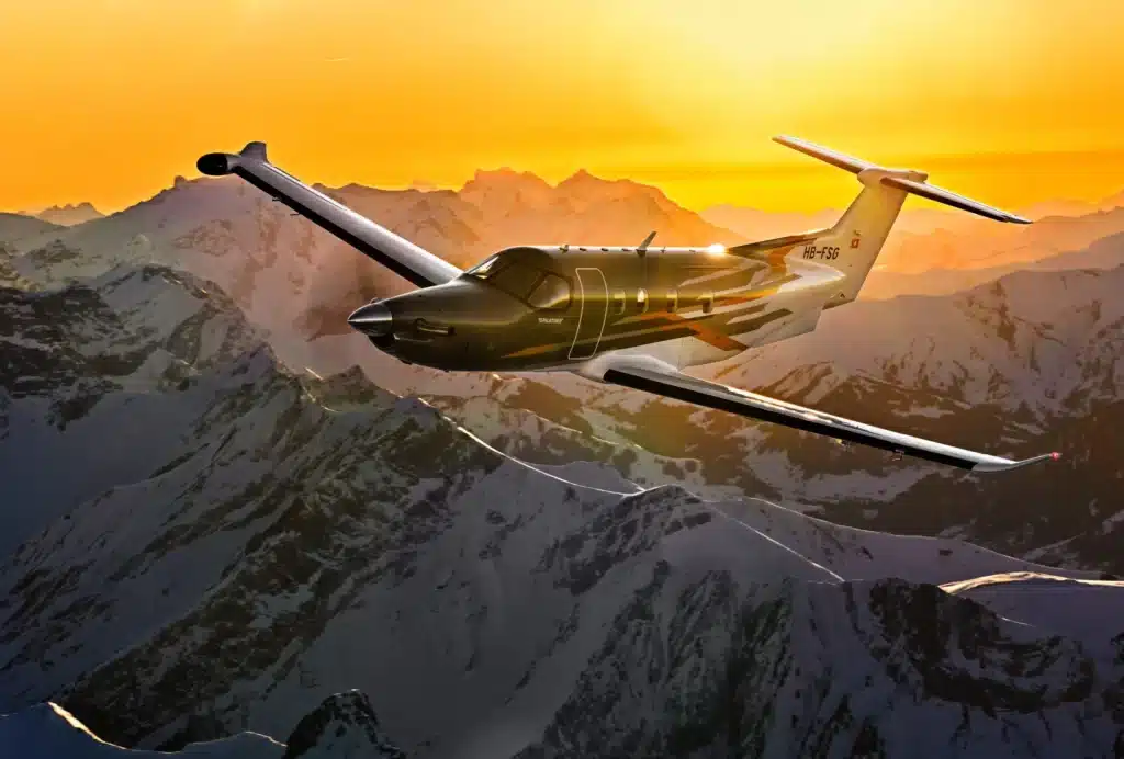 PC-12 aircraft flying over snow-covered mountains during sunset with a vibrant sky.