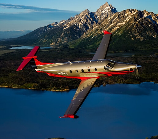 Pilatus PC-12 aircraft in gold and red livery flying over the sea with mountains in the background.