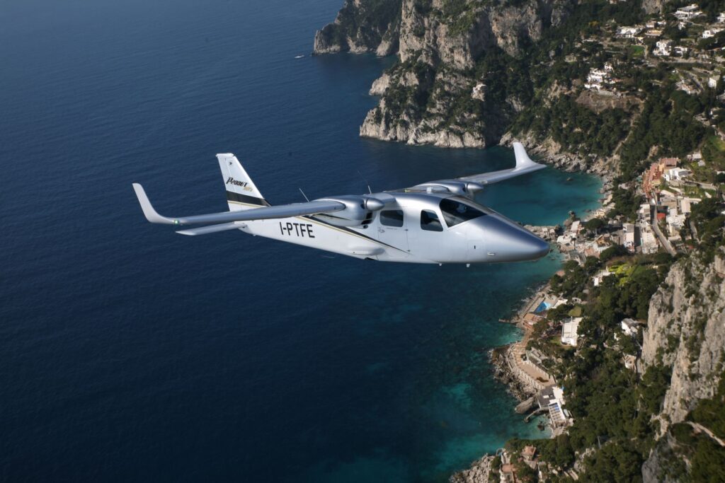 Tecnam P2006 aircraft in flight over a dark blue and turquoise sea with mountains in the background
