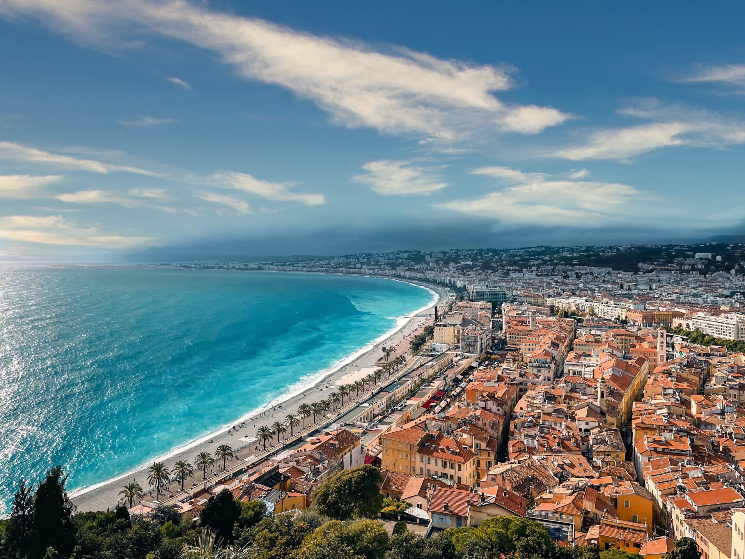 Aerial view of the coastline and city of Nice, France, featuring buildings and palm trees along the beach.