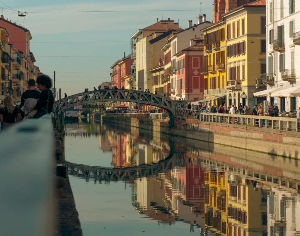 A canal reflecting colourful buildings and a bridge in a city setting with people walking along the banks.