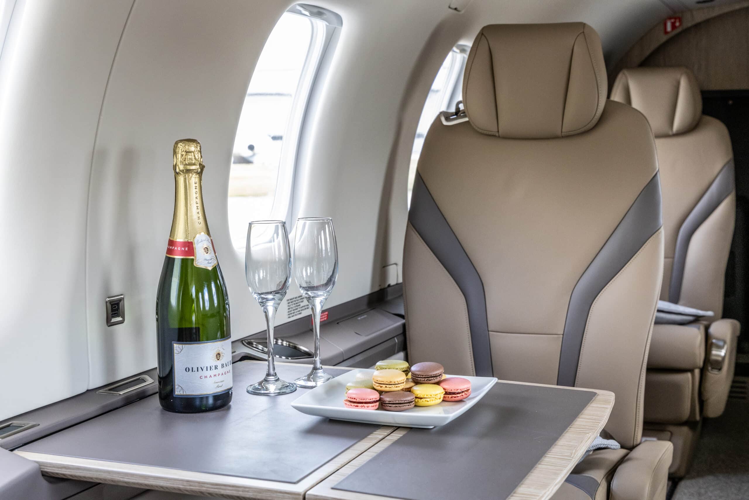 A bottle of champagne, two glasses, and a plate of macarons on a table inside a private jet cabin.